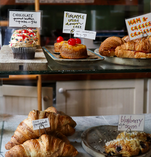 Assorted pastries and cakes for sale, including chocolate cupcake, pineapple upside-down cake, ham & cheese croissant, croissants, and blueberry bagels. Prices listed.