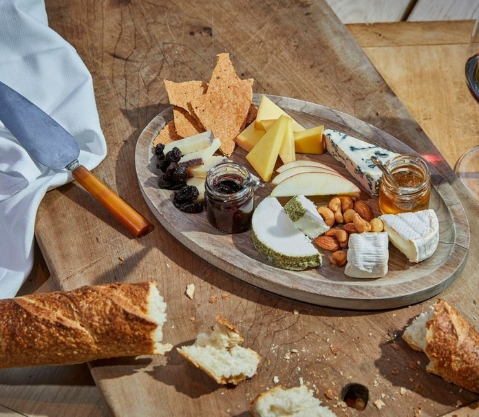 A cheese and charcuterie board with various cheeses, nuts, crackers, and preserves on a wooden board with a baguette and a wine glass nearby.