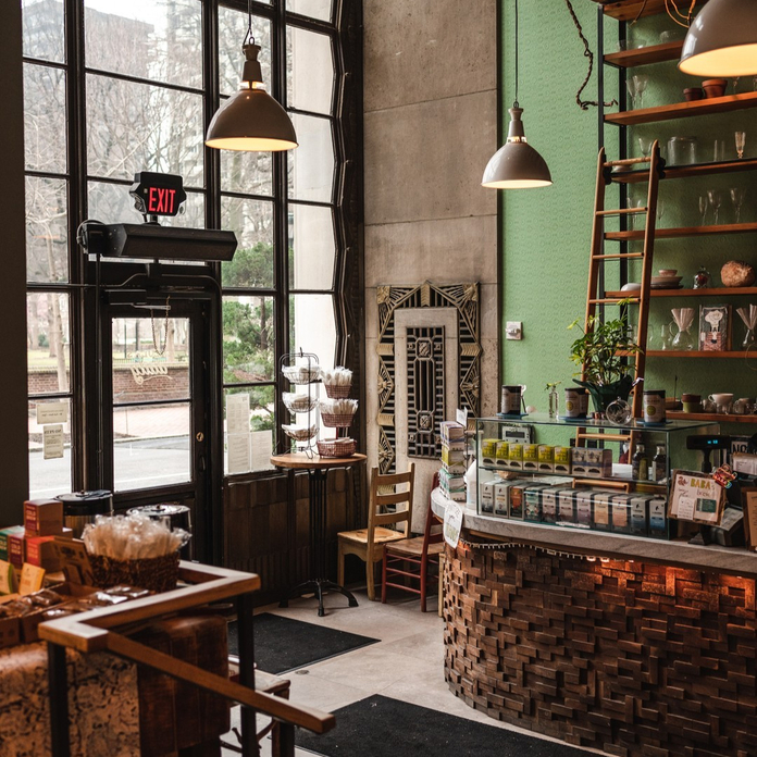 Modern restaurant interior with exposed brick, wooden furniture, large windows, and shelves displaying coffee items.